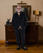 Man wearing navy corduroy suit standing in a room with a painting and lamp on a wall.