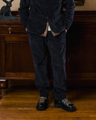 Person wearing a dark jacket, plaid shirt, and dark pants standing in a room with wooden furniture.