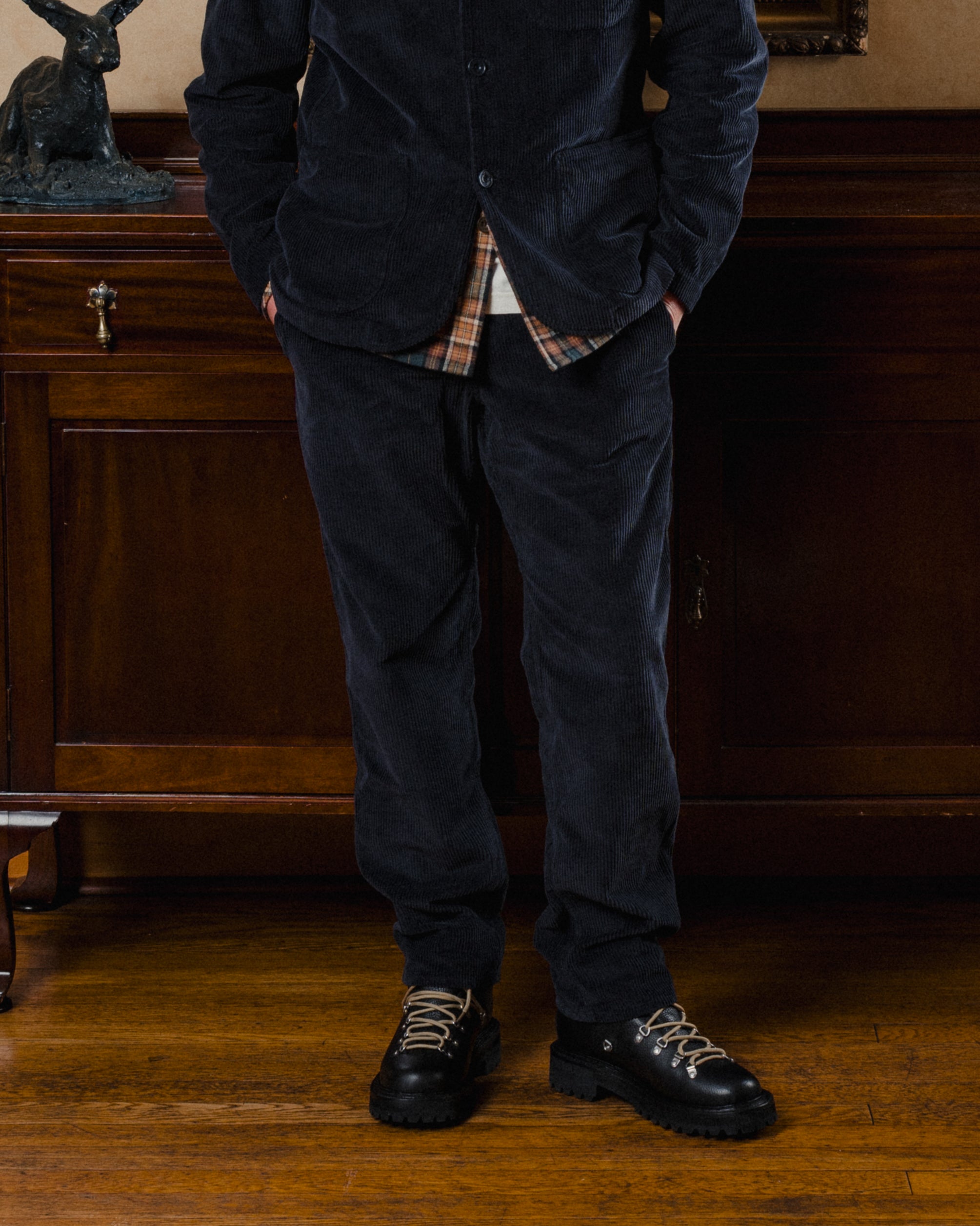 Person wearing a dark jacket, plaid shirt, and dark pants standing in a room with wooden furniture.