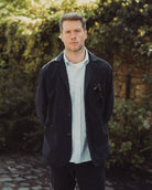 Man wearing a dark jacket over a light shirt standing outdoors with greenery in the background