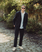 Man in dark coat and sunglasses standing on a stone path with greenery in the background