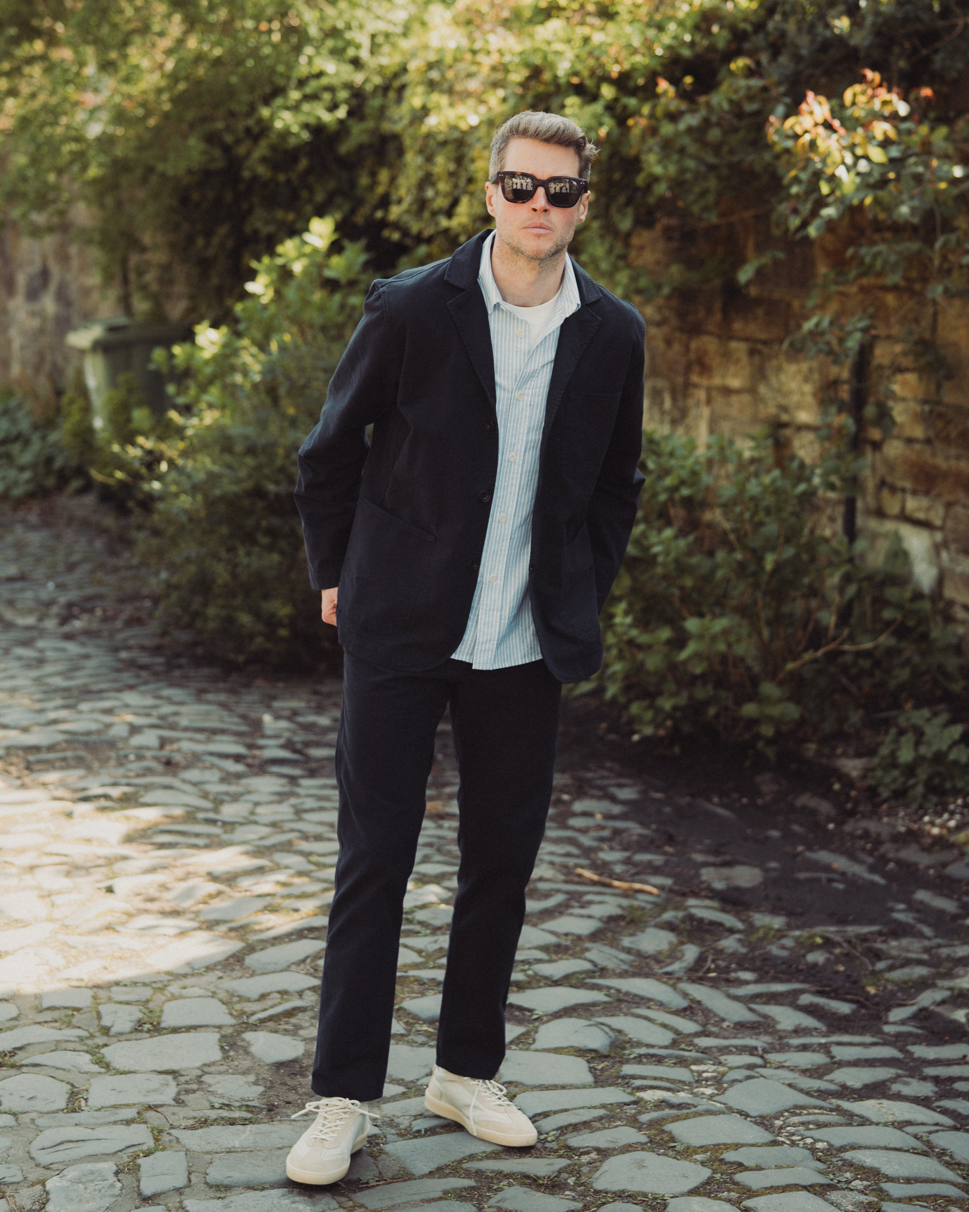 Man in dark coat and sunglasses standing on a stone path with greenery in the background