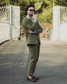 Man in a green suit standing on a pathway with white gates and greenery in the background