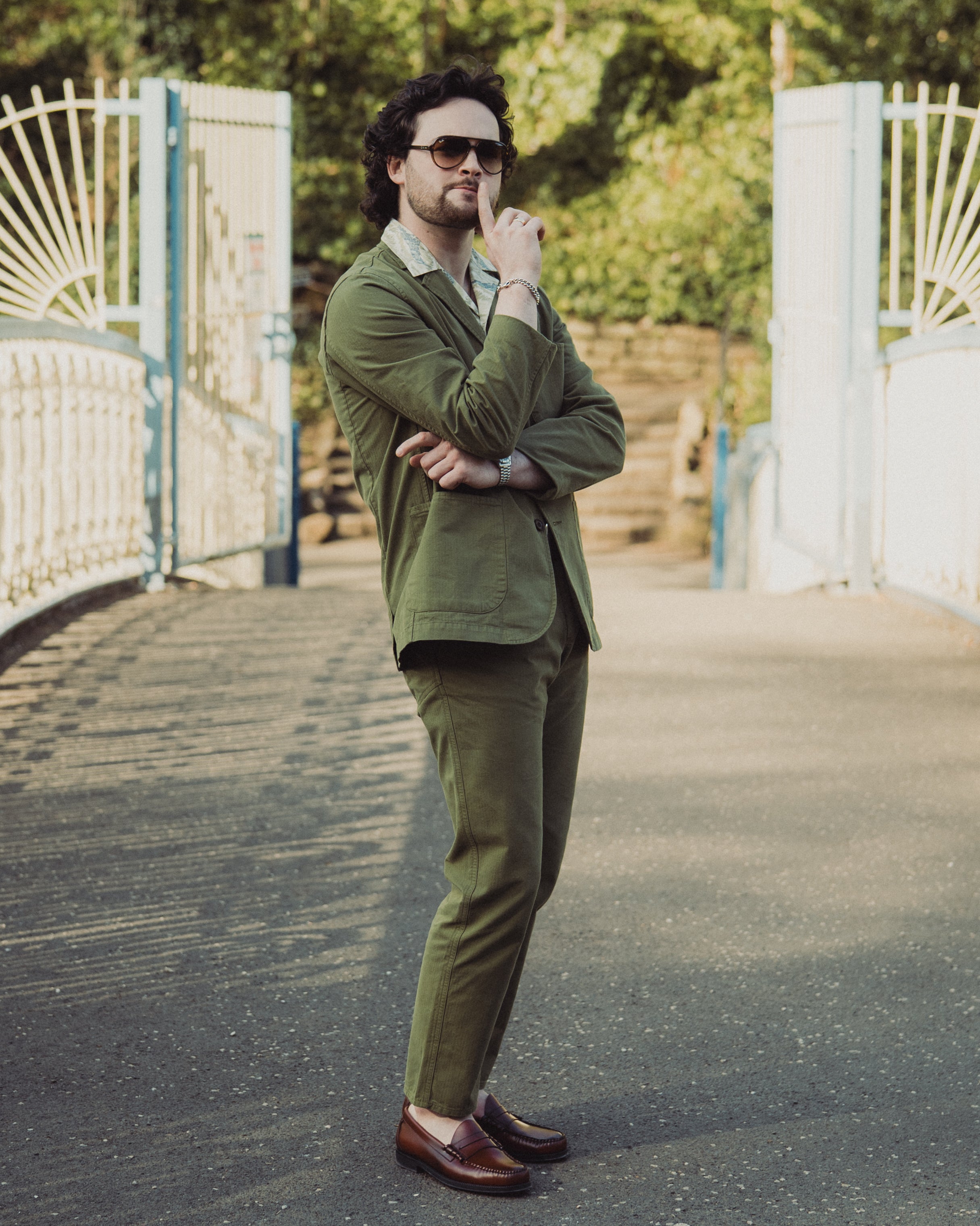 Man in a green suit standing on a pathway with white gates and greenery in the background