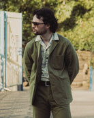 Man wearing a green jacket and sunglasses outdoors with greenery in the background