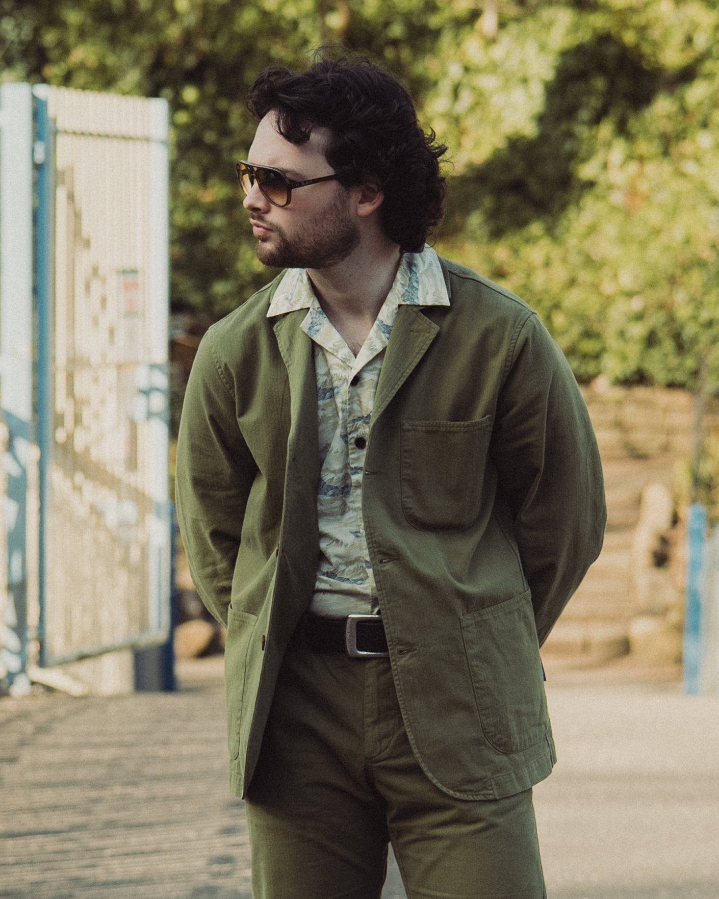 Man wearing a green jacket and sunglasses outdoors with greenery in the background