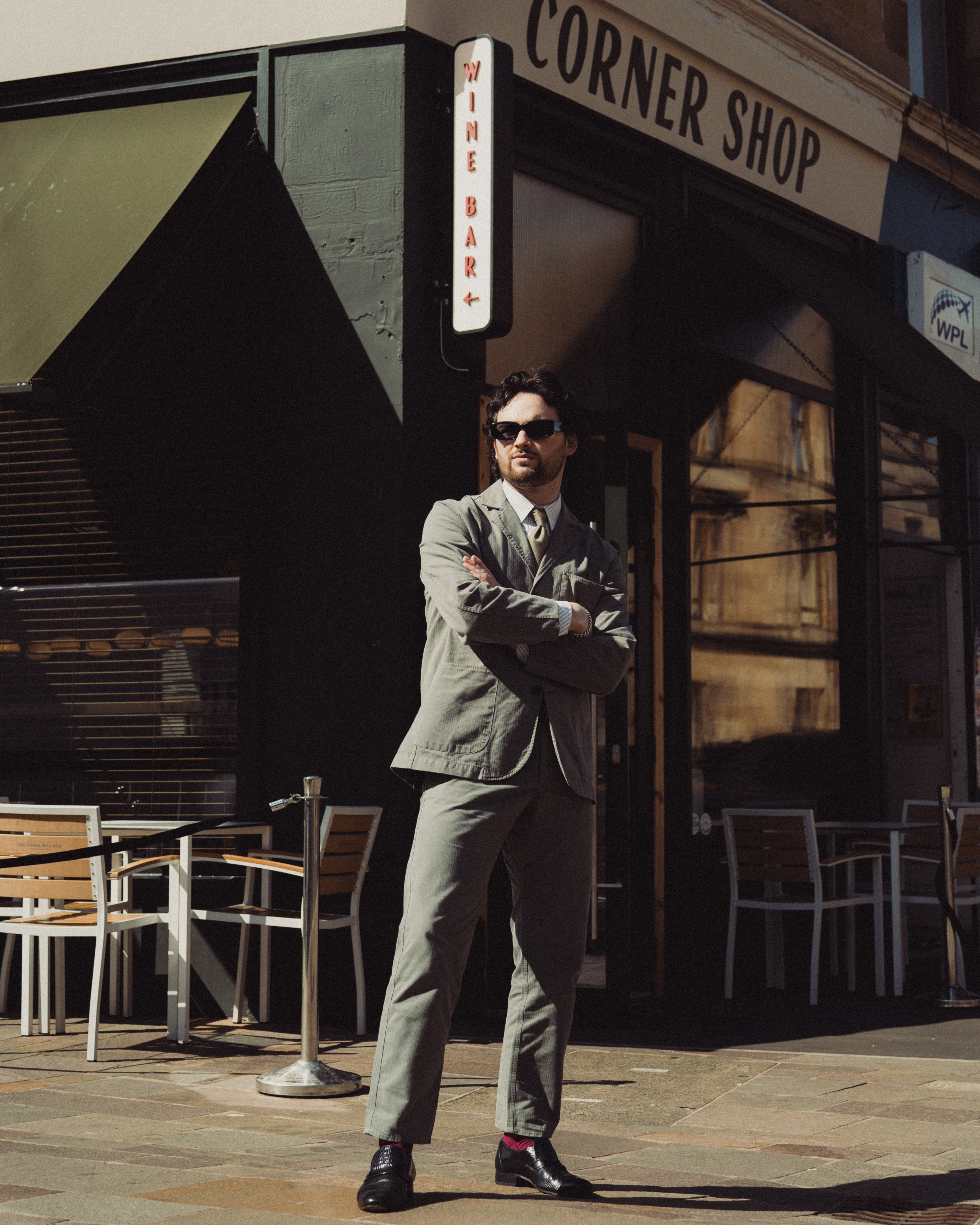 Man in a suit standing outside a corner shop with tables and chairs.