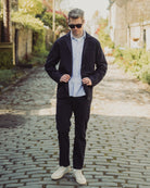 Man wearing a dark jacket, light shirt, and white sneakers on a cobblestone street.