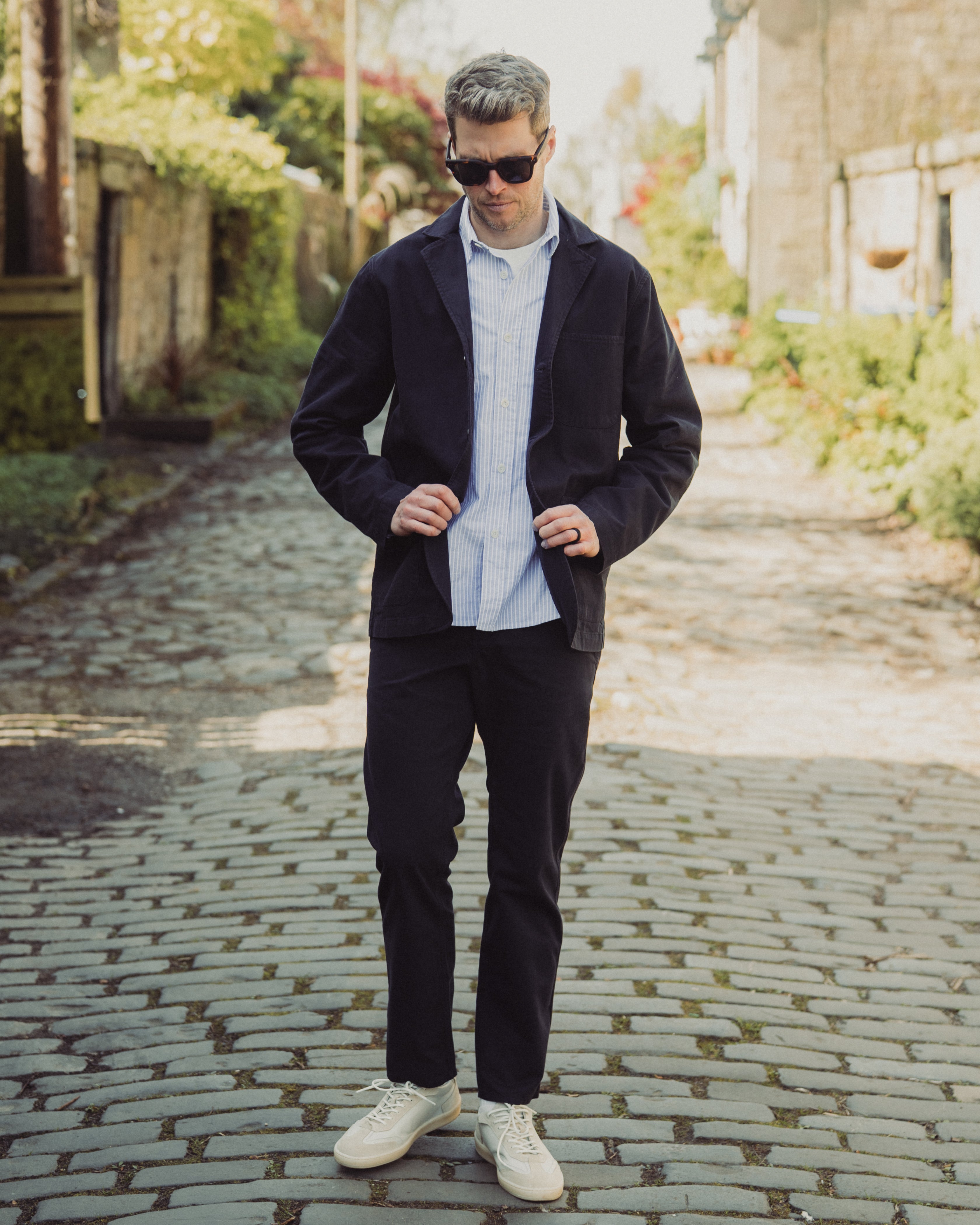 Man wearing a dark jacket, light shirt, and white sneakers on a cobblestone street.