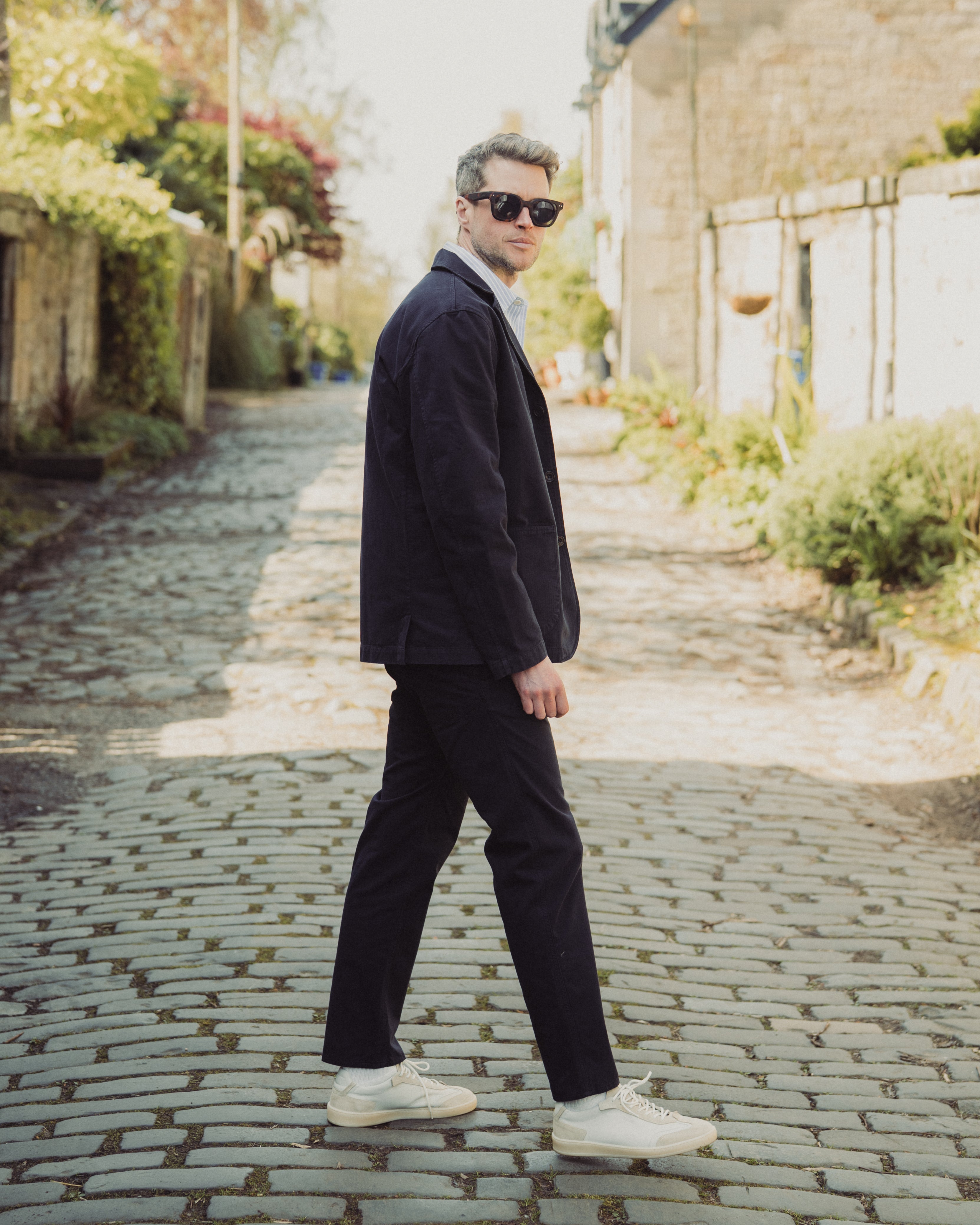 Man in a suit walking on a cobblestone street with greenery and a building in the background