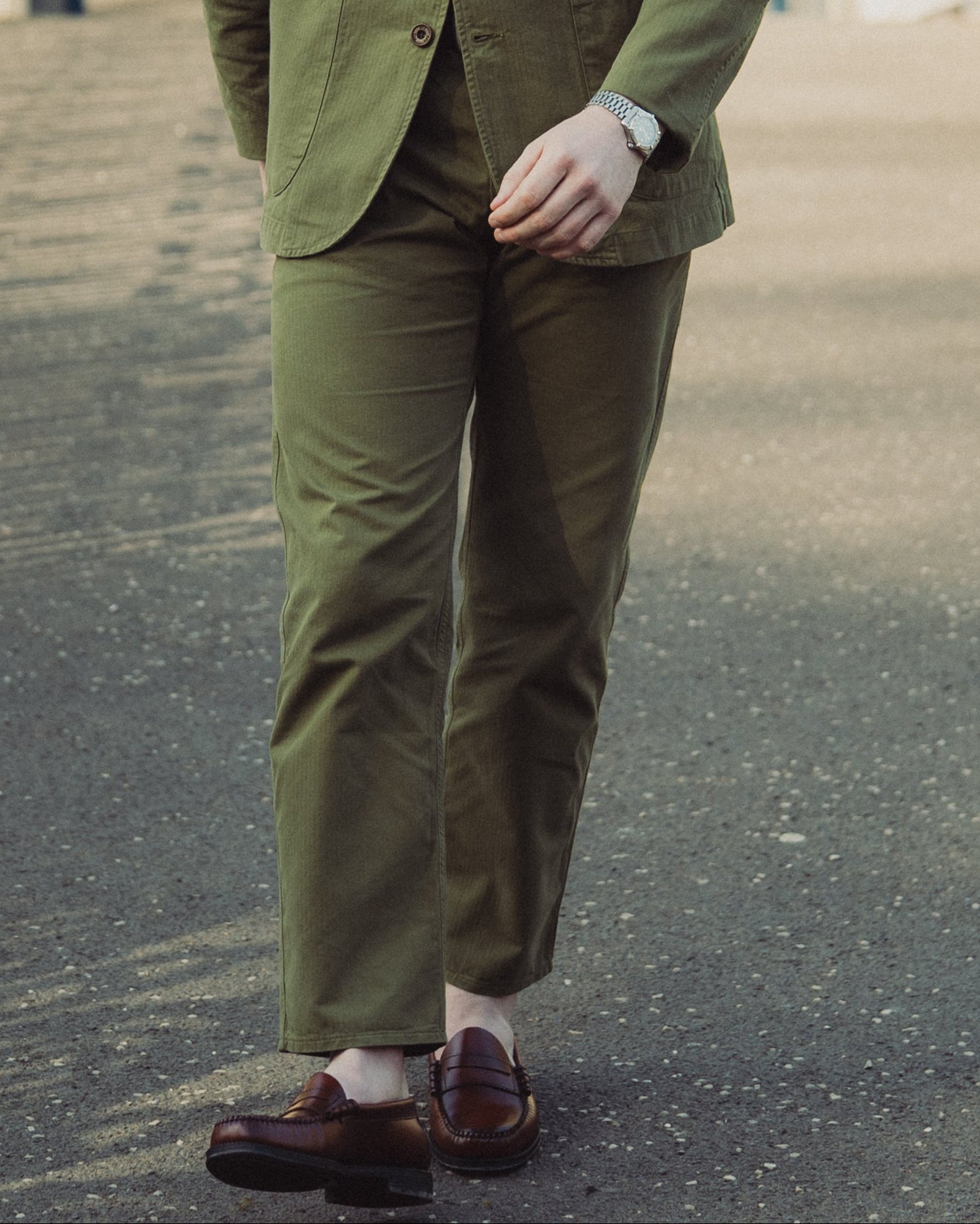 Man in a green jacket and pants standing on a paved path with a white gate in the background.