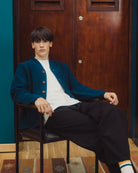 A young man sits in a black leather chair in front of a dark wood door, wearing a teal corduroy button-up cardigan over a white t-shirt, black trousers, and striped socks.