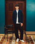 A man stands in a room wearing a dark navy blue cardigan over a white t-shirt, black pants, and white sneakers. The background features a dark wood door, teal walls, a black chair, and a patterned rug.