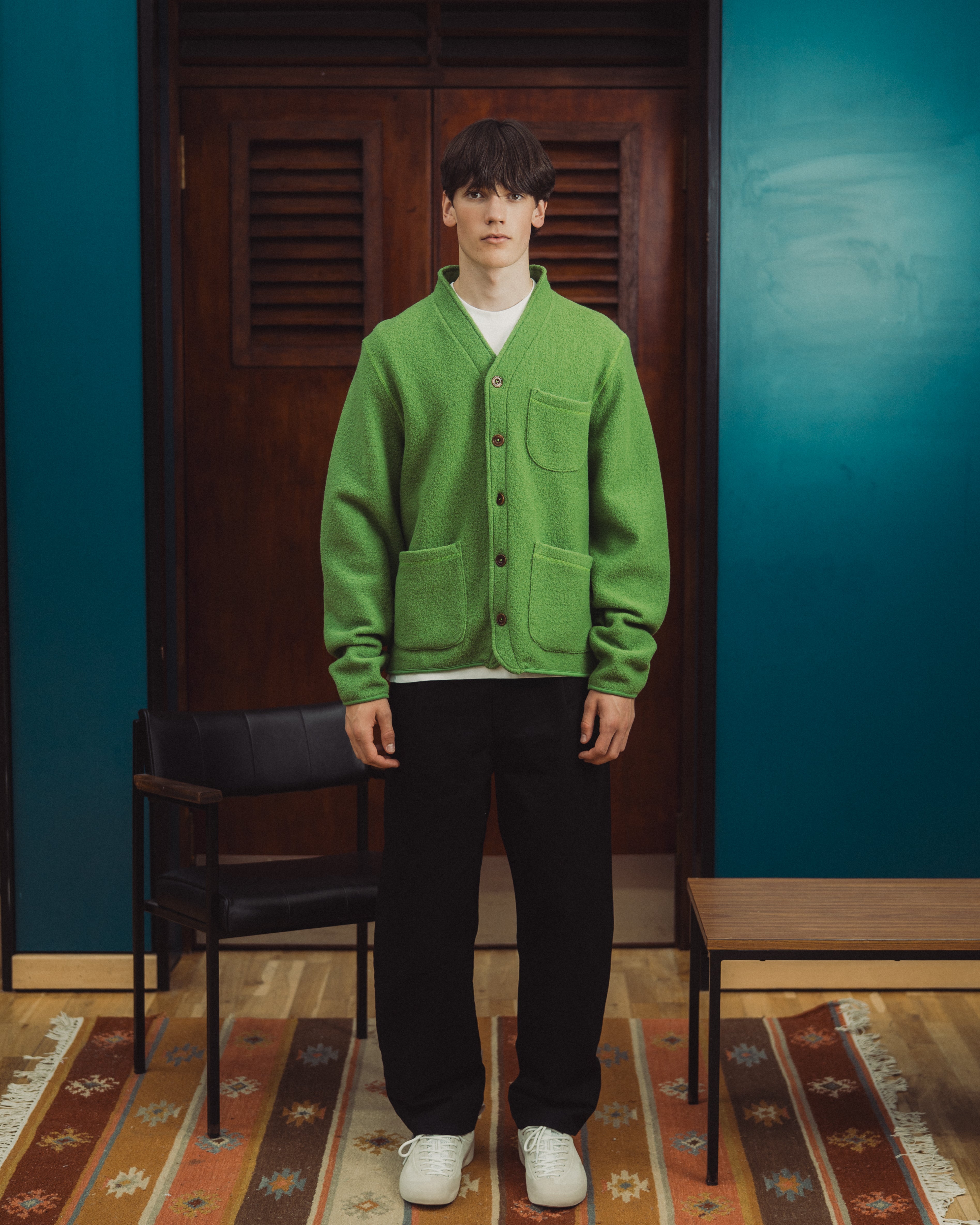 A man stands in a room wearing a bright green button-front cardigan over a white shirt, black pants, and white sneakers. The background features a dark wooden door, teal-blue walls, a black chair, and a patterned rug.