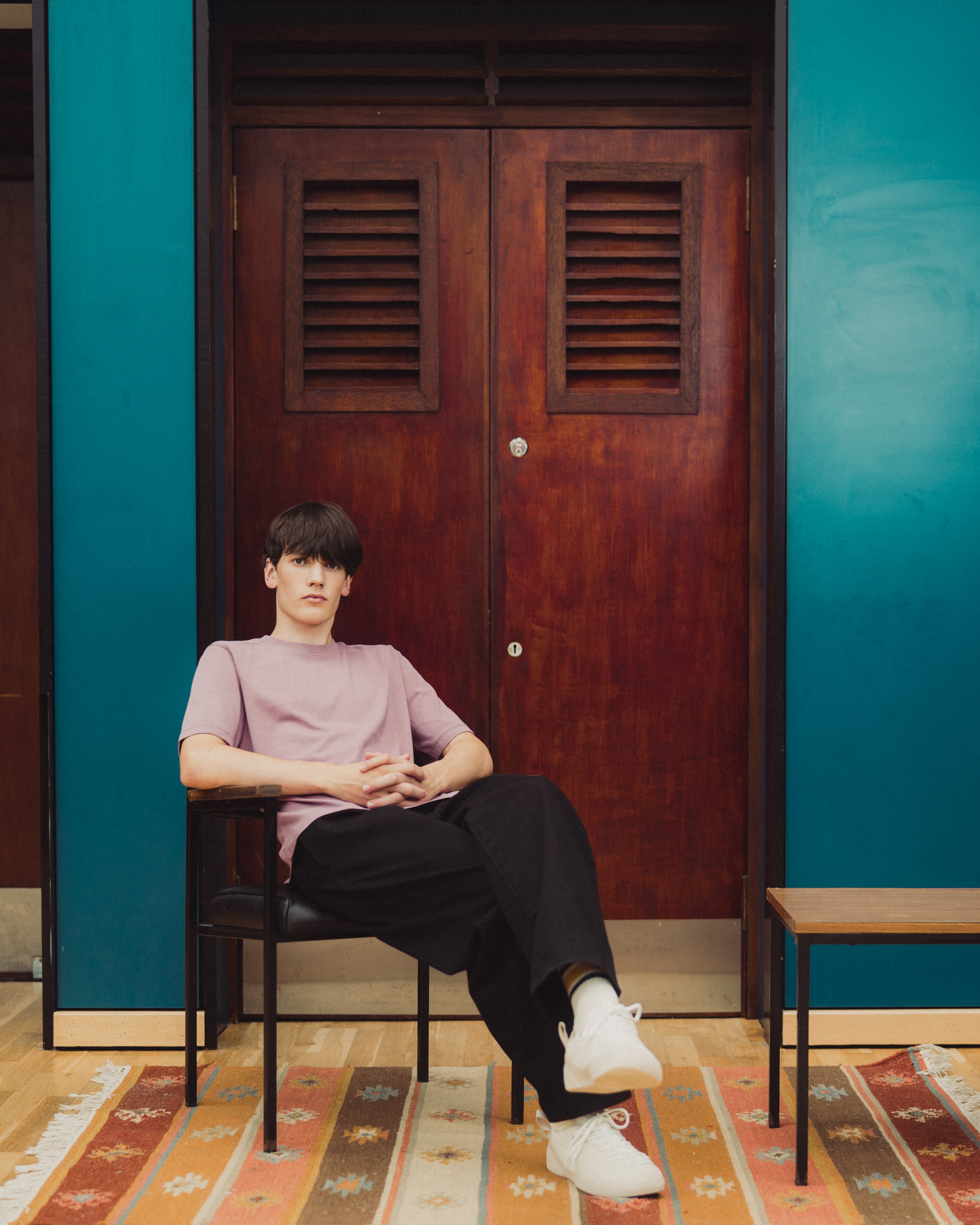 A man sits in a black chair in front of dark wooden doors, wearing a light pink t-shirt, black pants, and white sneakers. The room features teal blue walls and a patterned rug.