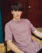 A young man wearing a dusty mauve-purple short-sleeve crewneck t-shirt, seated in front of a dark wood door.