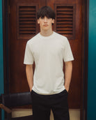 A young man wearing a plain white crewneck t-shirt and black pants, standing in front of a dark brown wooden door with louvered panels.