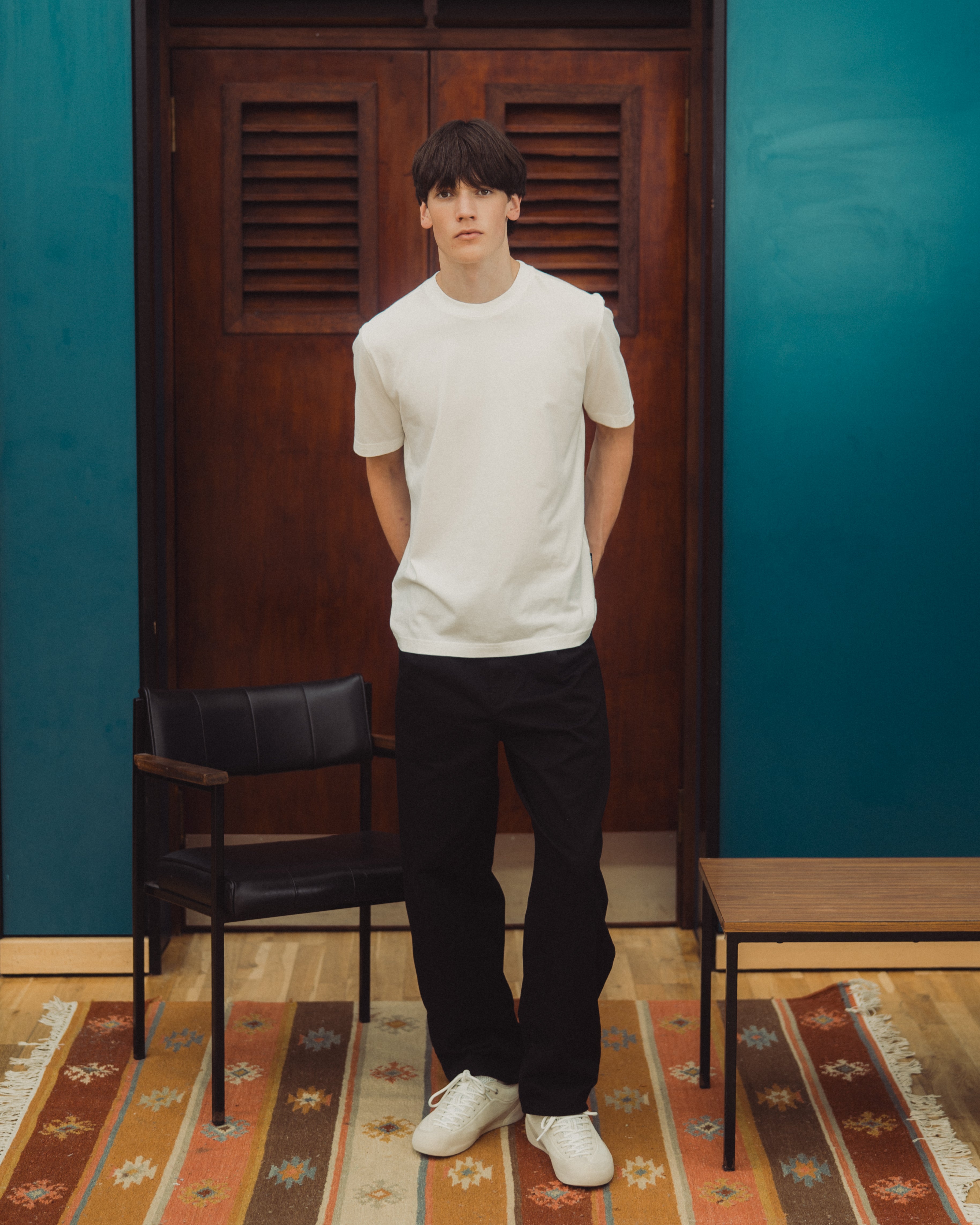 A man wearing a white crew-neck t-shirt, black pants, and white sneakers stands in front of a dark wooden door in a room with teal walls, a patterned rug, and a black chair.