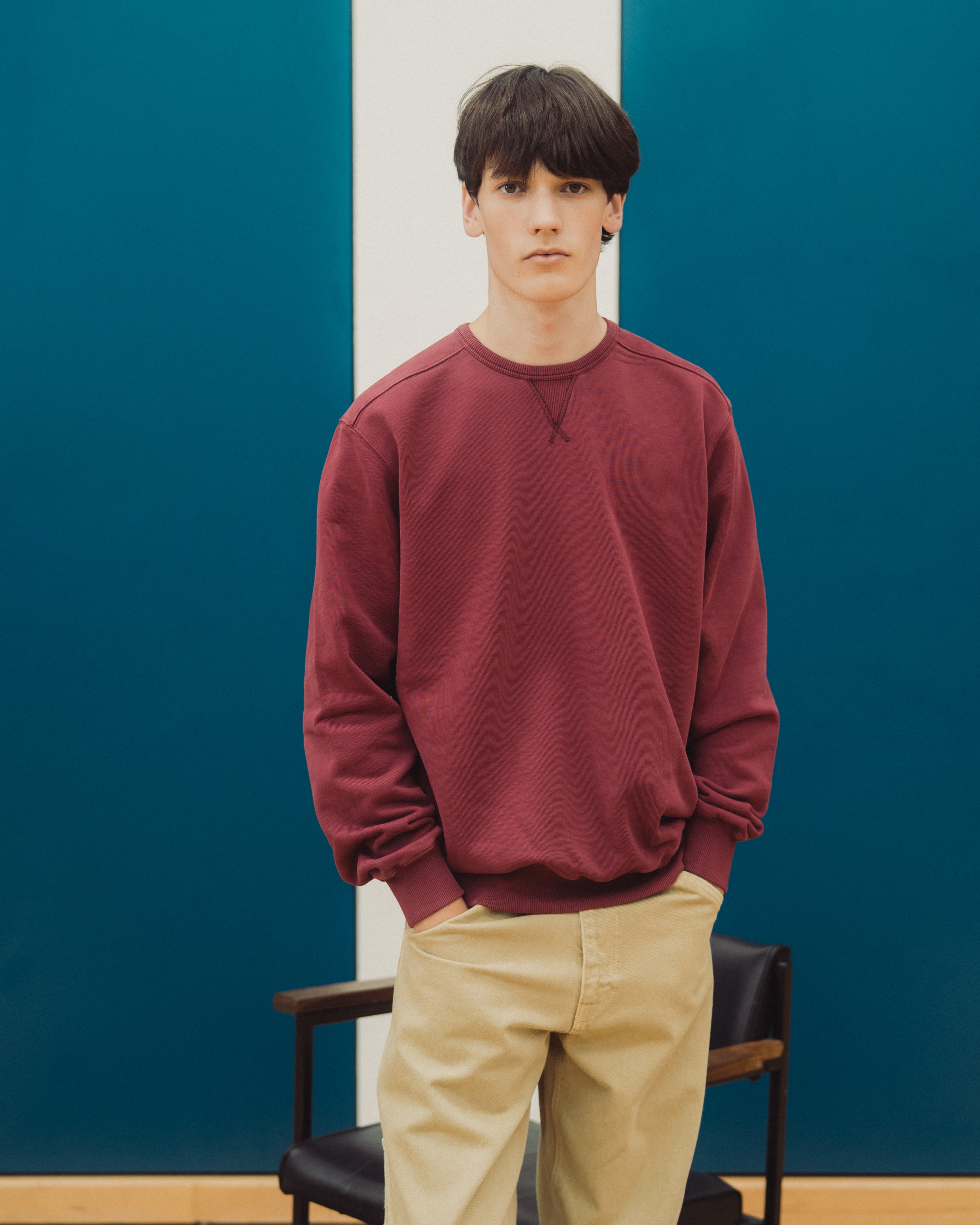 A young man wearing a crewneck sweatshirt in burgundy and beige pants, standing in front of a blue and white paneled wall.