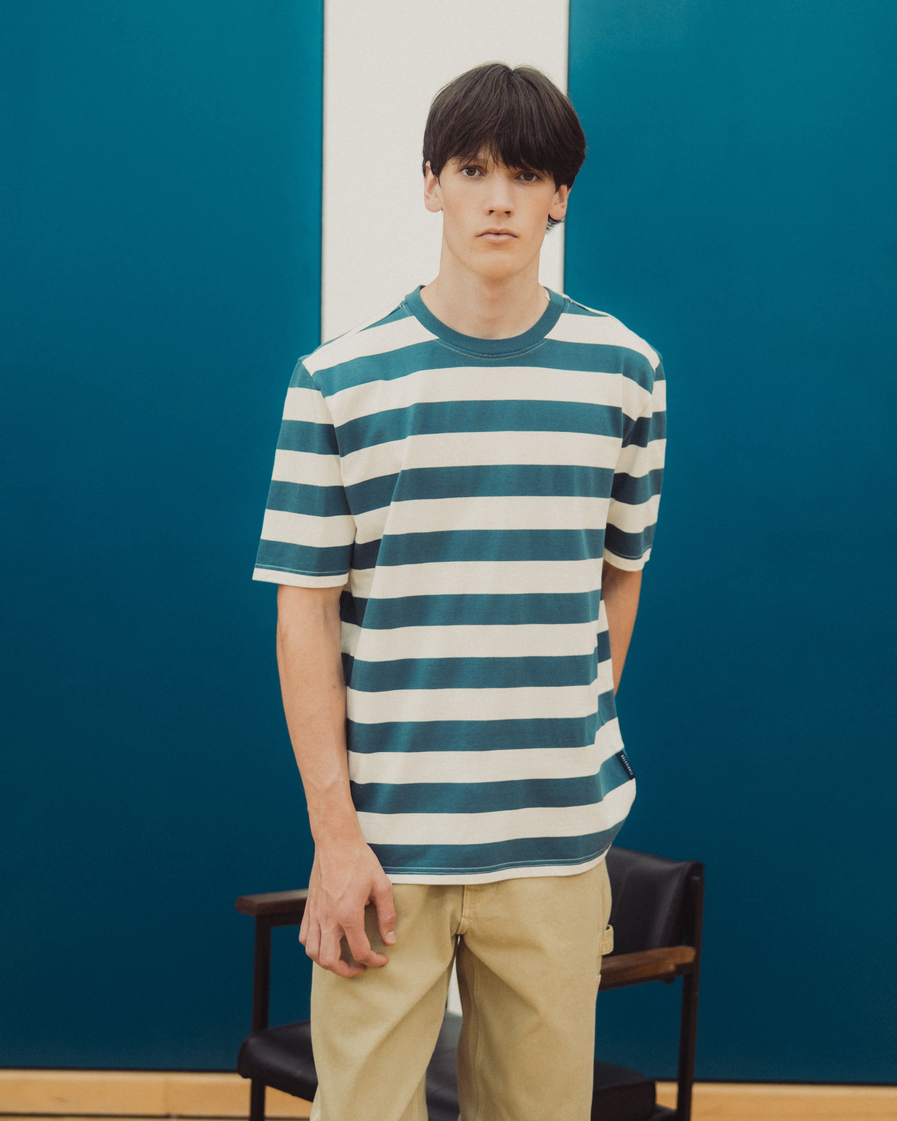 A young man wearing a teal and white horizontally striped short-sleeve t-shirt and khaki pants, standing in front of a blue and white paneled wall.