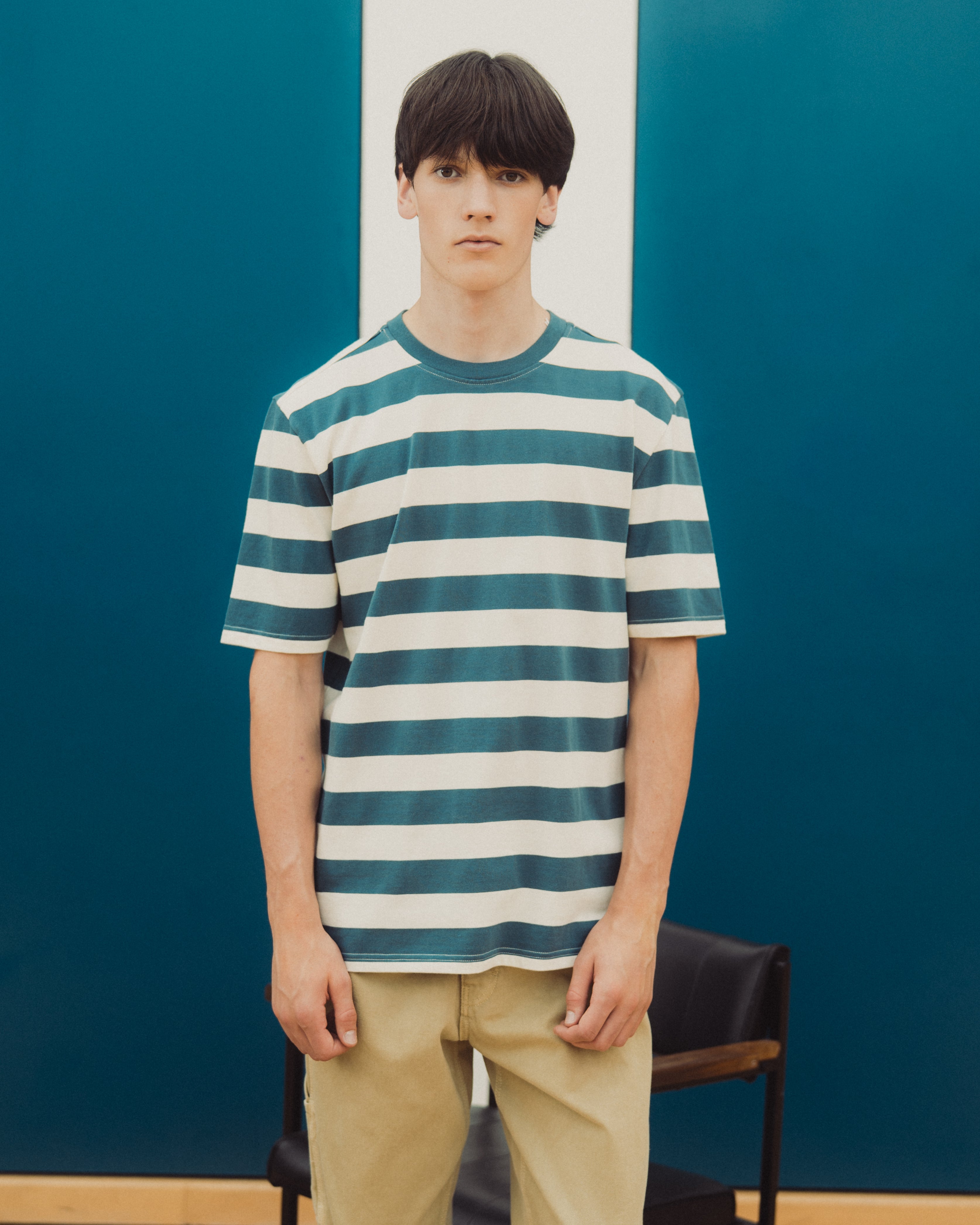 A young man wearing a teal and white horizontally striped t-shirt and khaki pants, standing in front of a dark teal wall with a white vertical stripe.
