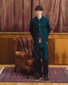 A young man stands in a room with dark brown velvet curtains, wood paneling, and a brown leather armchair, wearing a dark teal denim jacket and matching dark teal jeans over a pink shirt and dark sneakers.