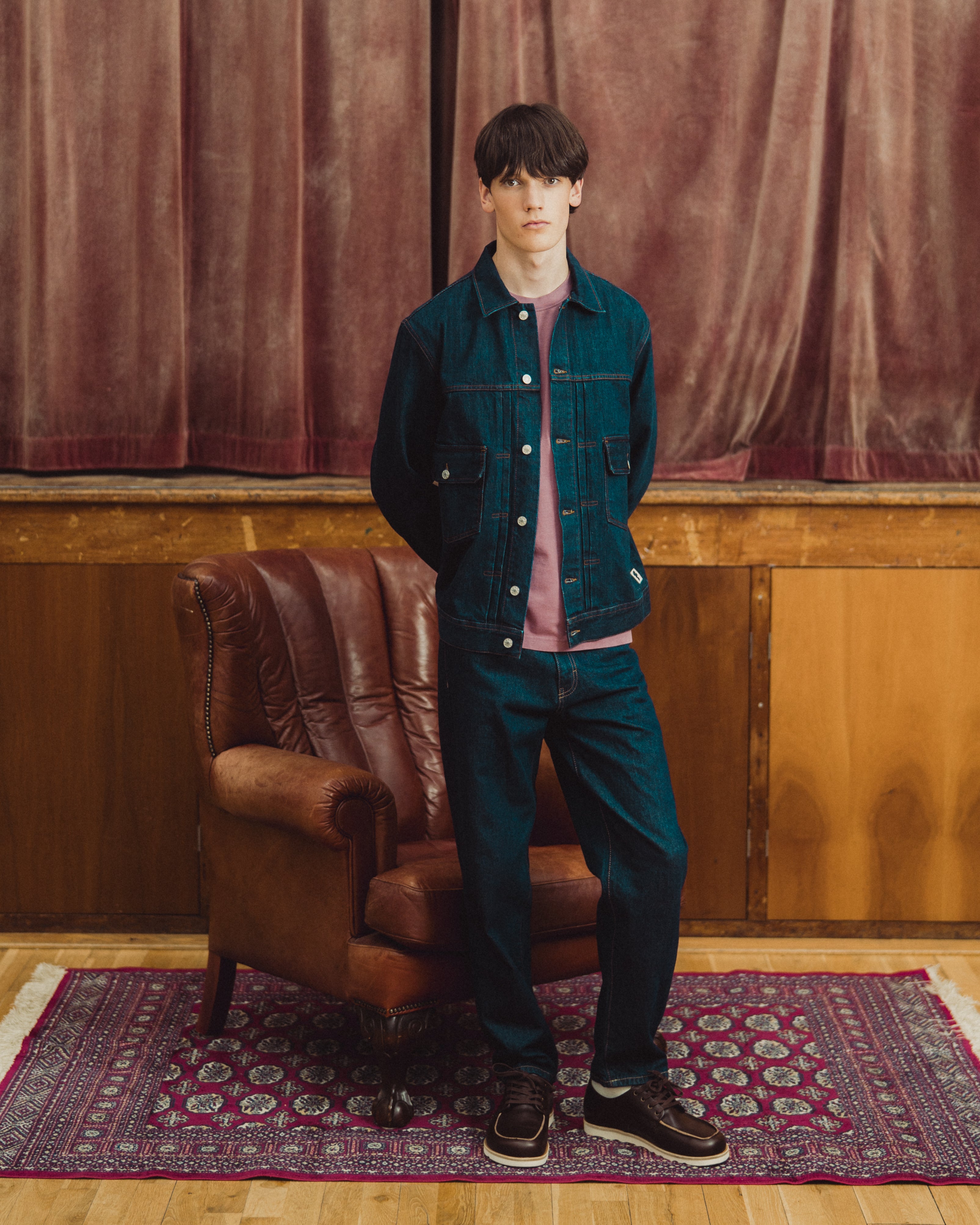 A model stands in a studio setting wearing a dark teal denim jacket over a dusty pink t-shirt, matching dark teal denim jeans, and dark brown sneakers. He is posed next to a brown leather armchair on a patterned rug in front of a wooden paneled wall and a rust-colored velvet curtain.