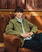 A young man sits in a brown leather armchair wearing an olive green utility jacket over a light blue and white striped shirt, paired with dark blue jeans.