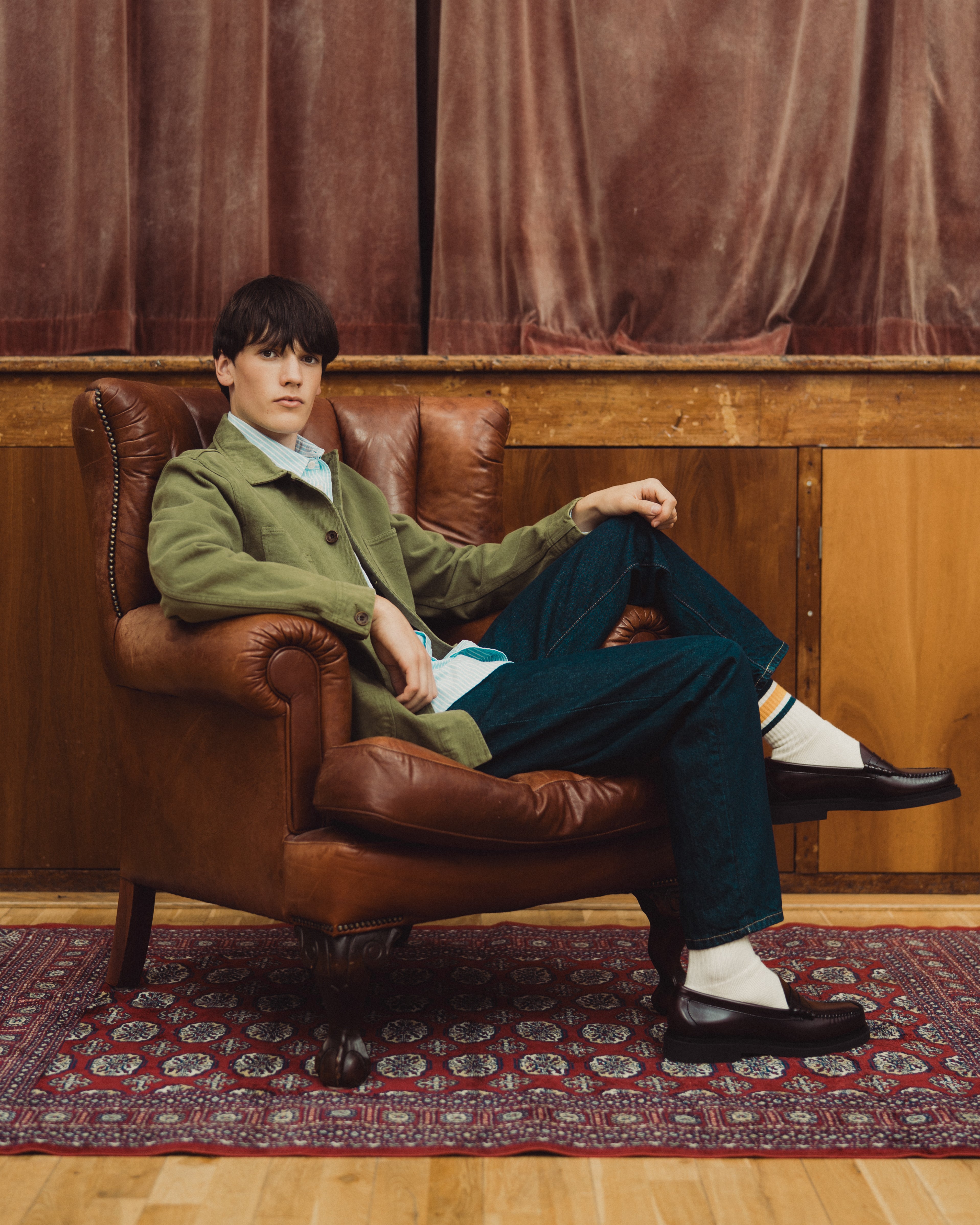 A man sits in a brown leather armchair wearing a green blazer, light blue shirt, dark teal trousers, white socks, and black loafers, posed in front of a wooden panel wall and brown curtain.