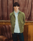 A young man wearing an olive green utility jacket over a light blue and white striped button-down shirt, paired with dark blue jeans, standing in front of a brown leather armchair and a brown velvet curtain.