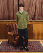 A young man stands in a room wearing an olive green corduroy jacket over a light blue collared shirt, paired with dark trousers and black loafers. He is positioned in front of a brown leather armchair and a patterned red rug, with brown velvet curtains and wooden paneling in the background.