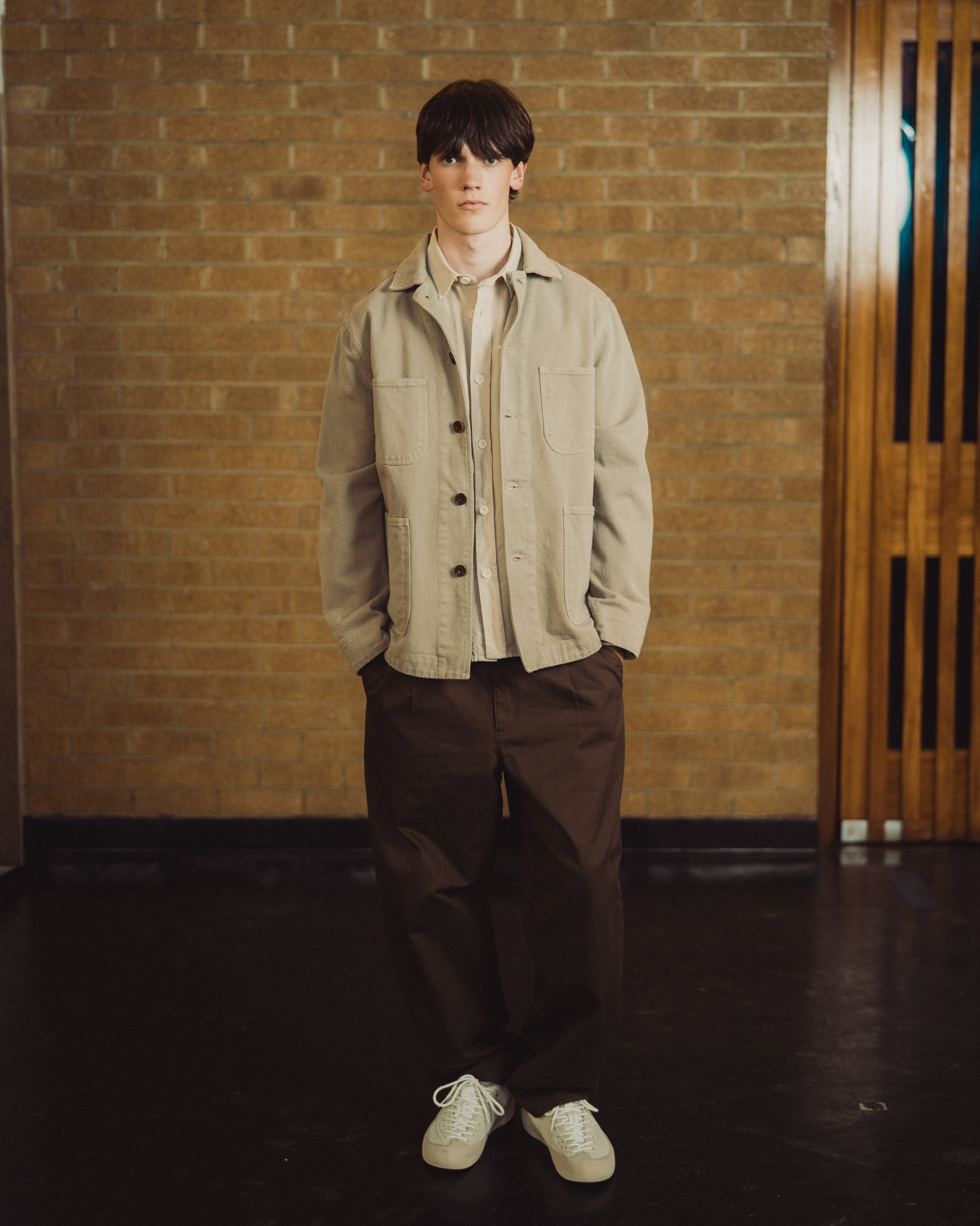 A young man stands in front of a brick wall wearing a light beige utility jacket over a cream shirt, dark brown trousers, and white sneakers.
