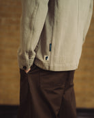 Close-up of a person wearing a light beige canvas utility jacket with a small embroidered logo patch and a vertical brand tab, paired with dark brown pants, set against a warm blurred brick wall background.