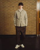 A young man stands in front of a brick wall wearing a beige utility jacket over a white collared shirt, dark brown trousers, and off-white sneakers.
