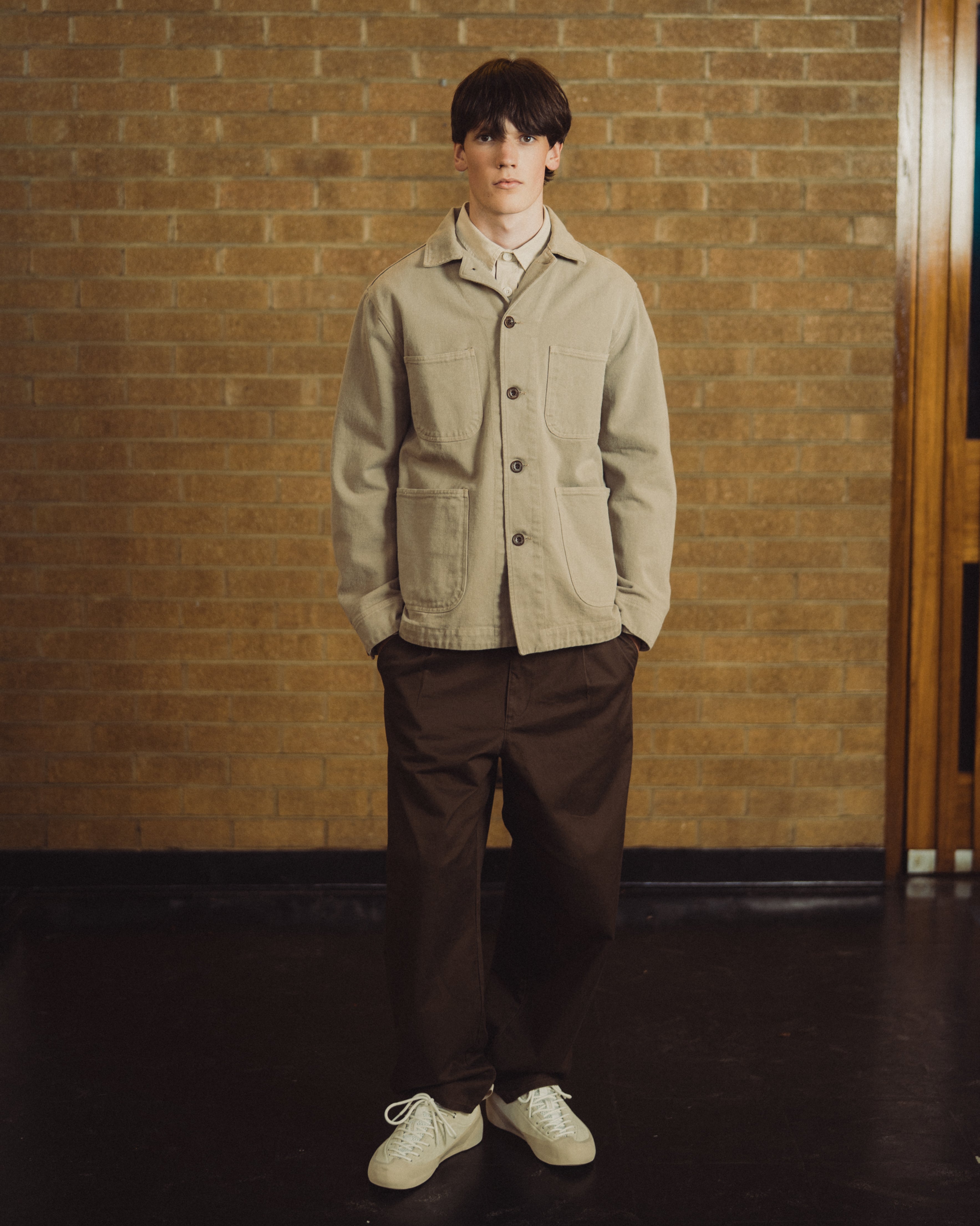 A young man stands in front of a brick wall wearing a beige utility jacket over a white collared shirt, dark brown trousers, and off-white sneakers.