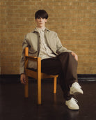 A young man sits on a wooden chair against a tan brick wall, wearing a light beige overshirt over a cream shirt, dark brown trousers, and white sneakers.