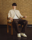 A man sits on a wooden chair in front of a tan brick wall, wearing a light beige overshirt over a white t-shirt, dark brown trousers, and off-white sneakers.