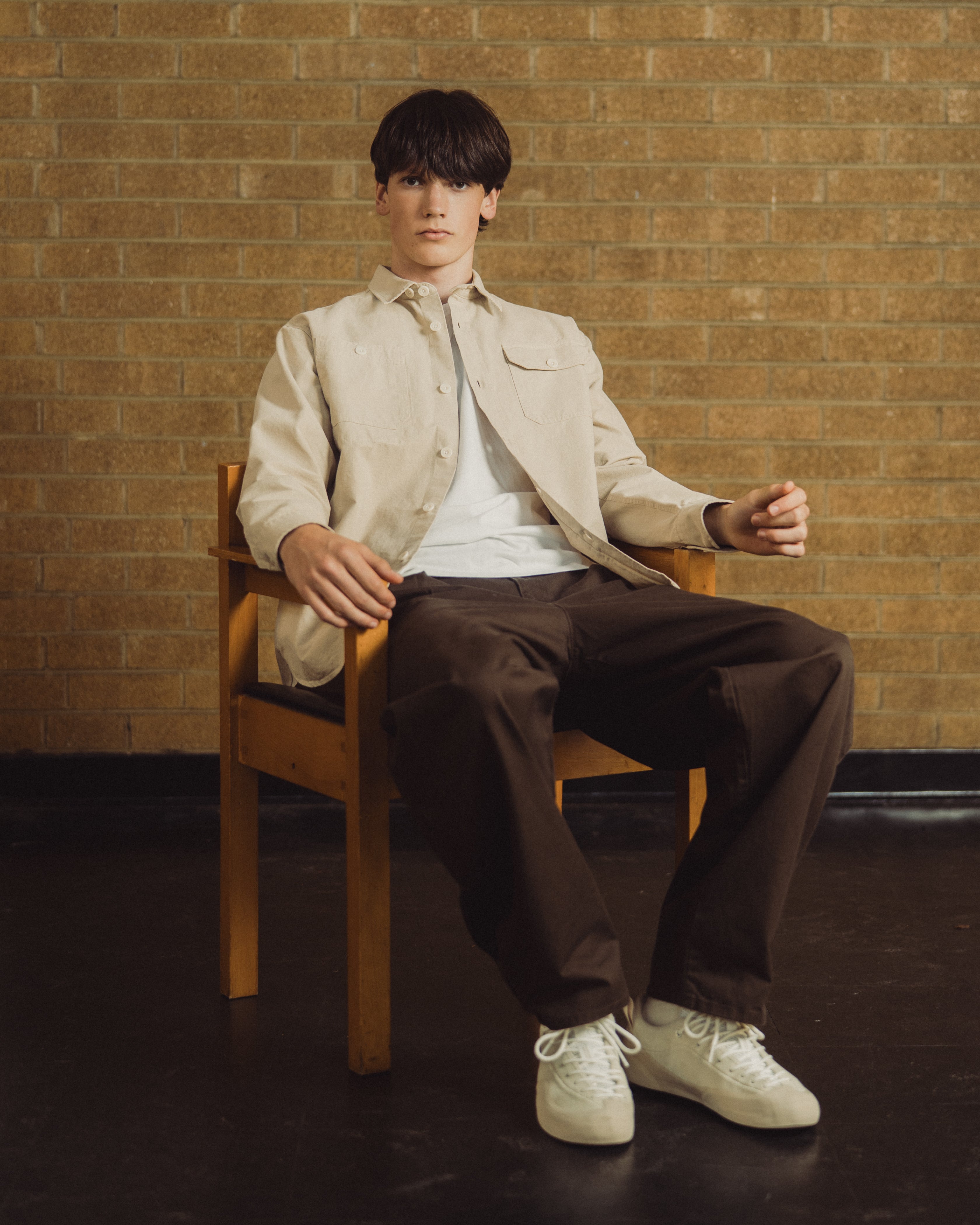 A man sits on a wooden chair in front of a tan brick wall, wearing a light beige overshirt over a white t-shirt, dark brown trousers, and off-white sneakers.