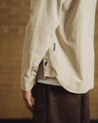 A man sits on a wooden chair in front of a tan brick wall, wearing a light beige overshirt over a white t-shirt, dark brown trousers, and off-white sneakers.