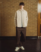 A man sits on a wooden chair in front of a tan brick wall, wearing a light beige overshirt over a white t-shirt, dark brown trousers, and off-white sneakers.