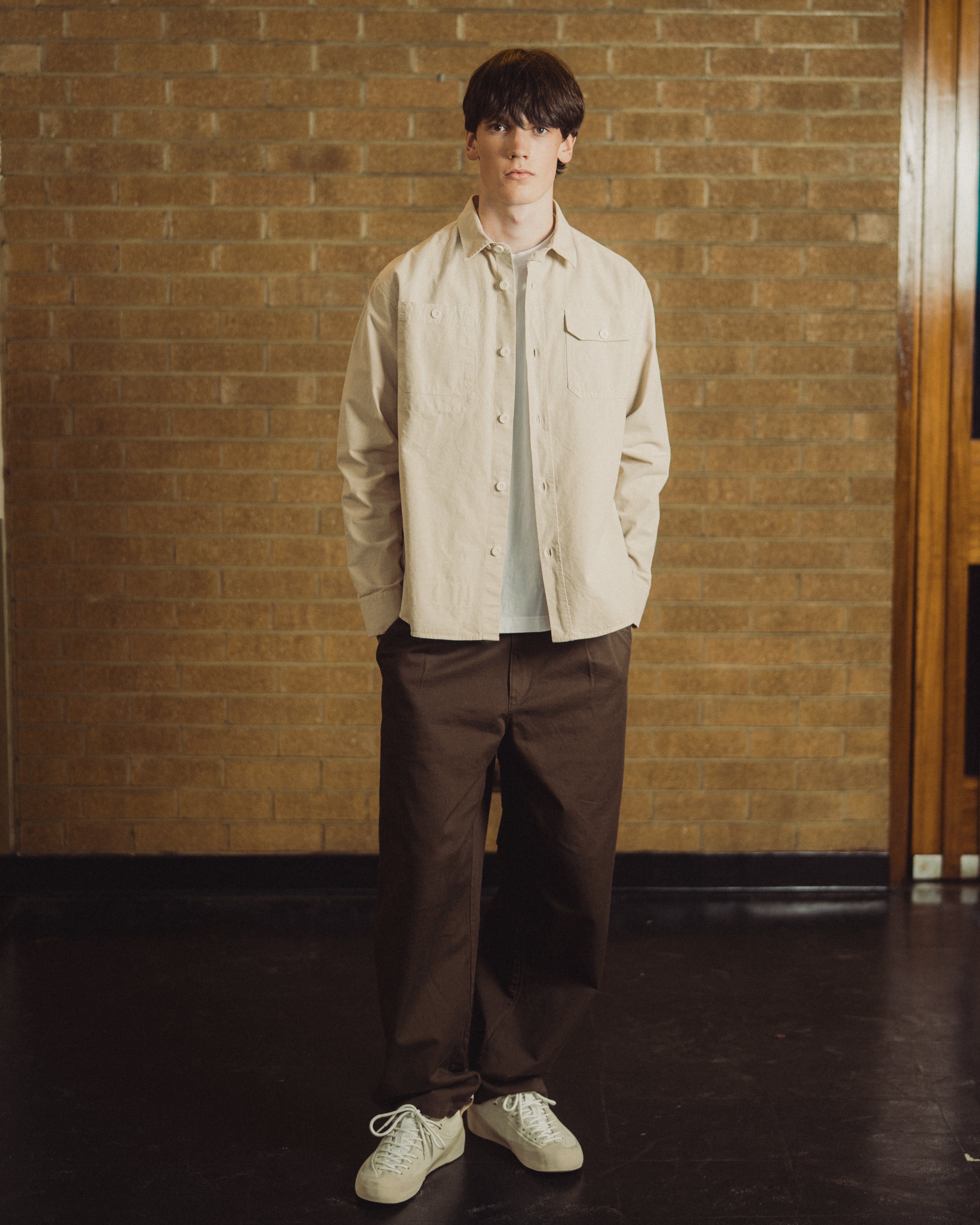 A man sits on a wooden chair in front of a tan brick wall, wearing a light beige overshirt over a white t-shirt, dark brown trousers, and off-white sneakers.