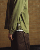 A person wearing an olive green button-up shirt and brown pants, shown from the side against a warm brick wall background.