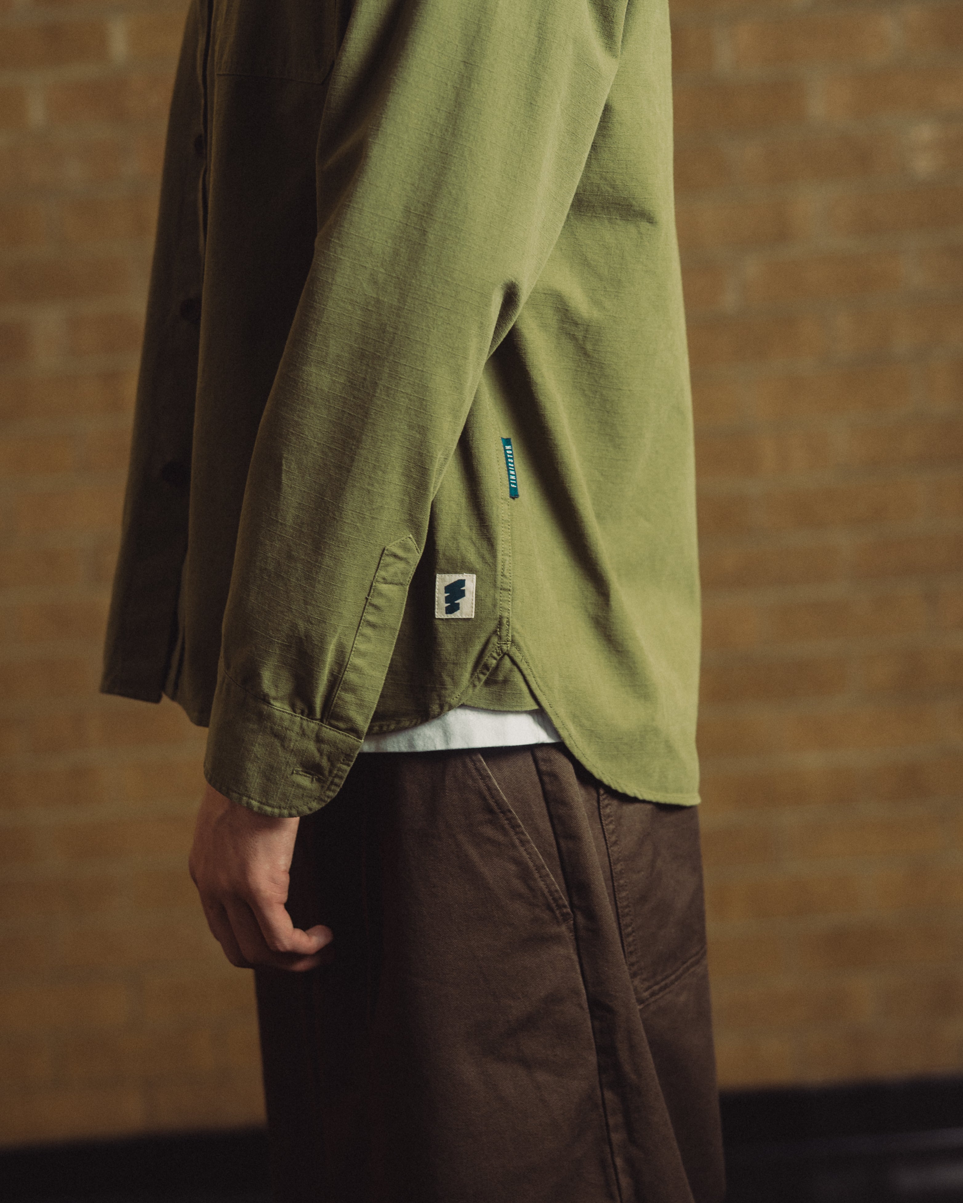 A person wearing an olive green button-up shirt and brown pants, shown from the side against a warm brick wall background.