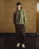 A young man stands in front of a tan brick wall wearing an olive green pullover hoodie, dark brown pants, and off-white sneakers.