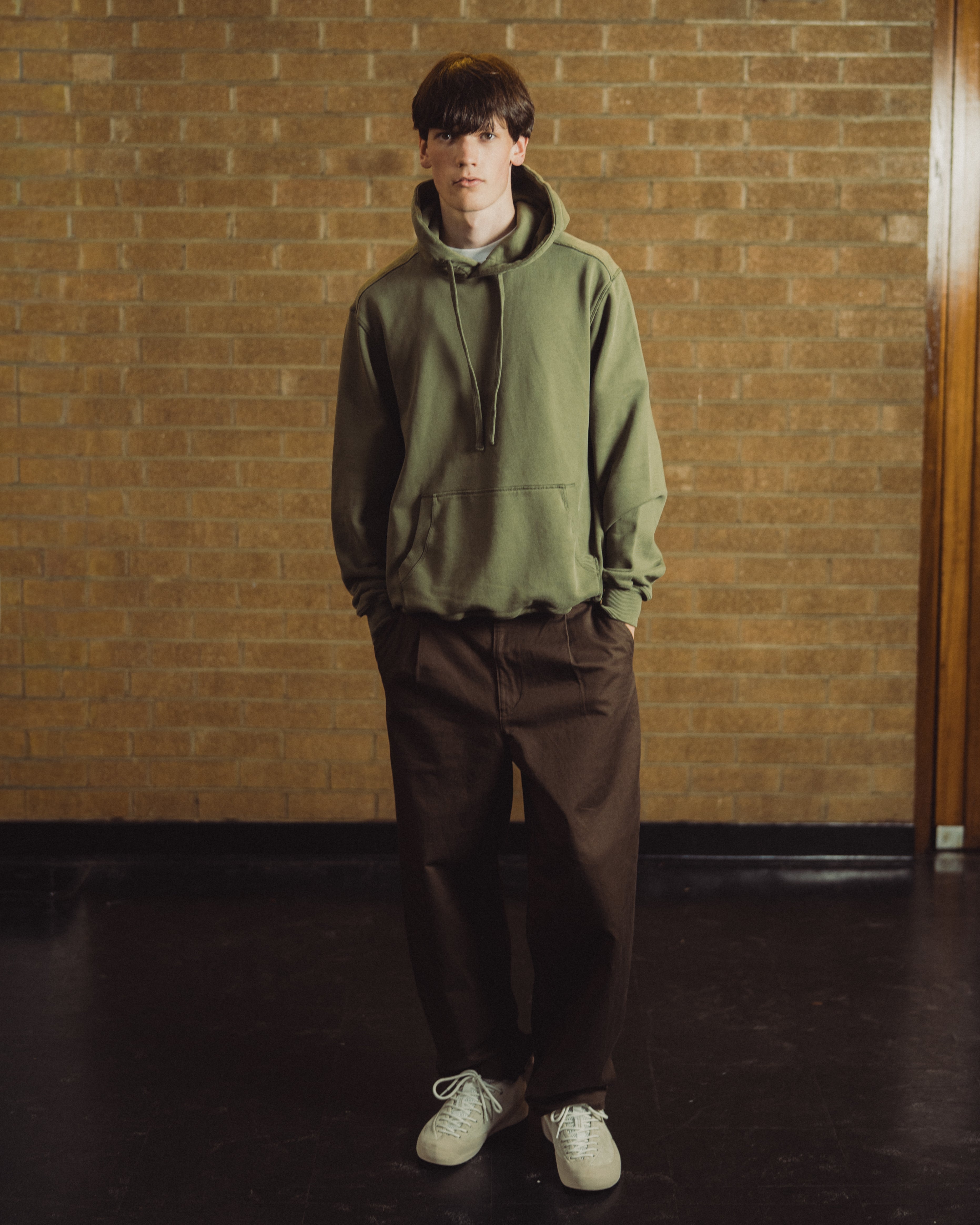 A young man stands in front of a tan brick wall wearing an olive green pullover hoodie, dark brown pants, and off-white sneakers.