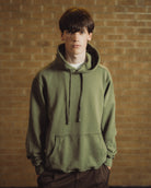 A young man wearing an olive green pullover hoodie with drawstrings and a kangaroo pocket, standing against a warm-toned brick wall background.