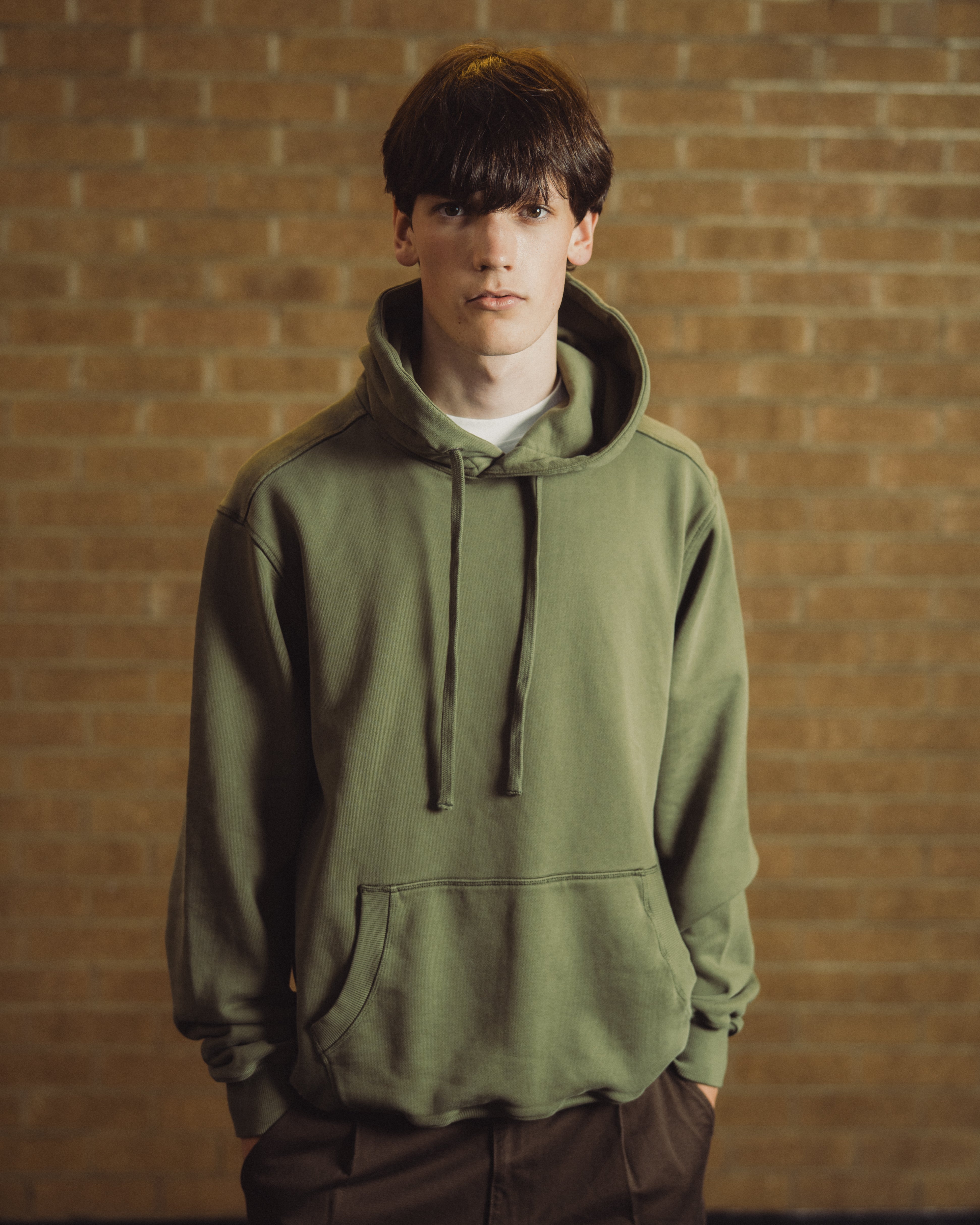 A young man wearing an olive green pullover hoodie with drawstrings and a kangaroo pocket, standing against a warm-toned brick wall background.