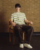 A young man sits on a wooden chair against a brick wall, wearing a light green and white horizontally striped t-shirt, dark brown pants, and white sneakers.