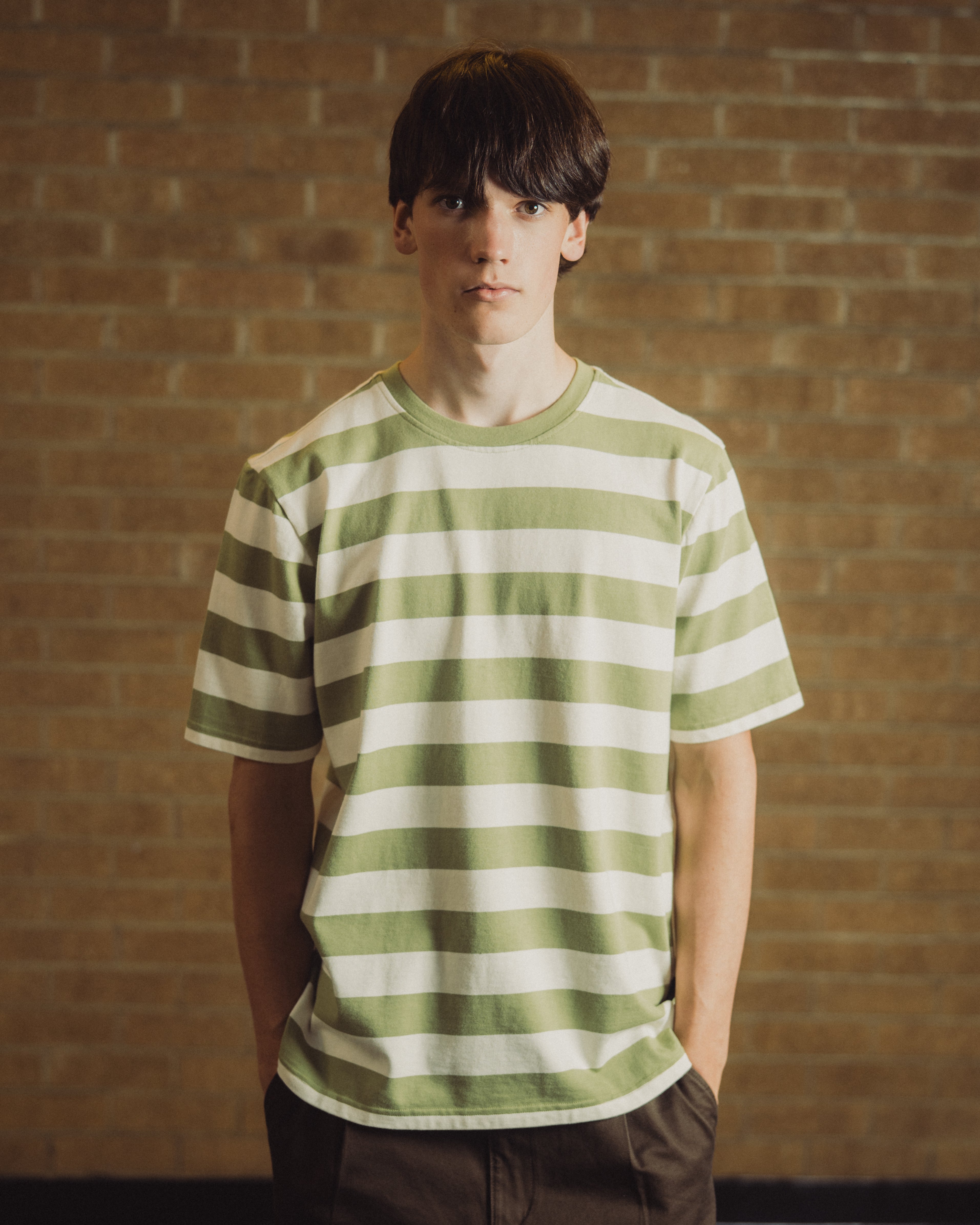 A young man wearing a green and white horizontally striped t-shirt, standing with hands in his pockets against a warm-toned brick wall background.