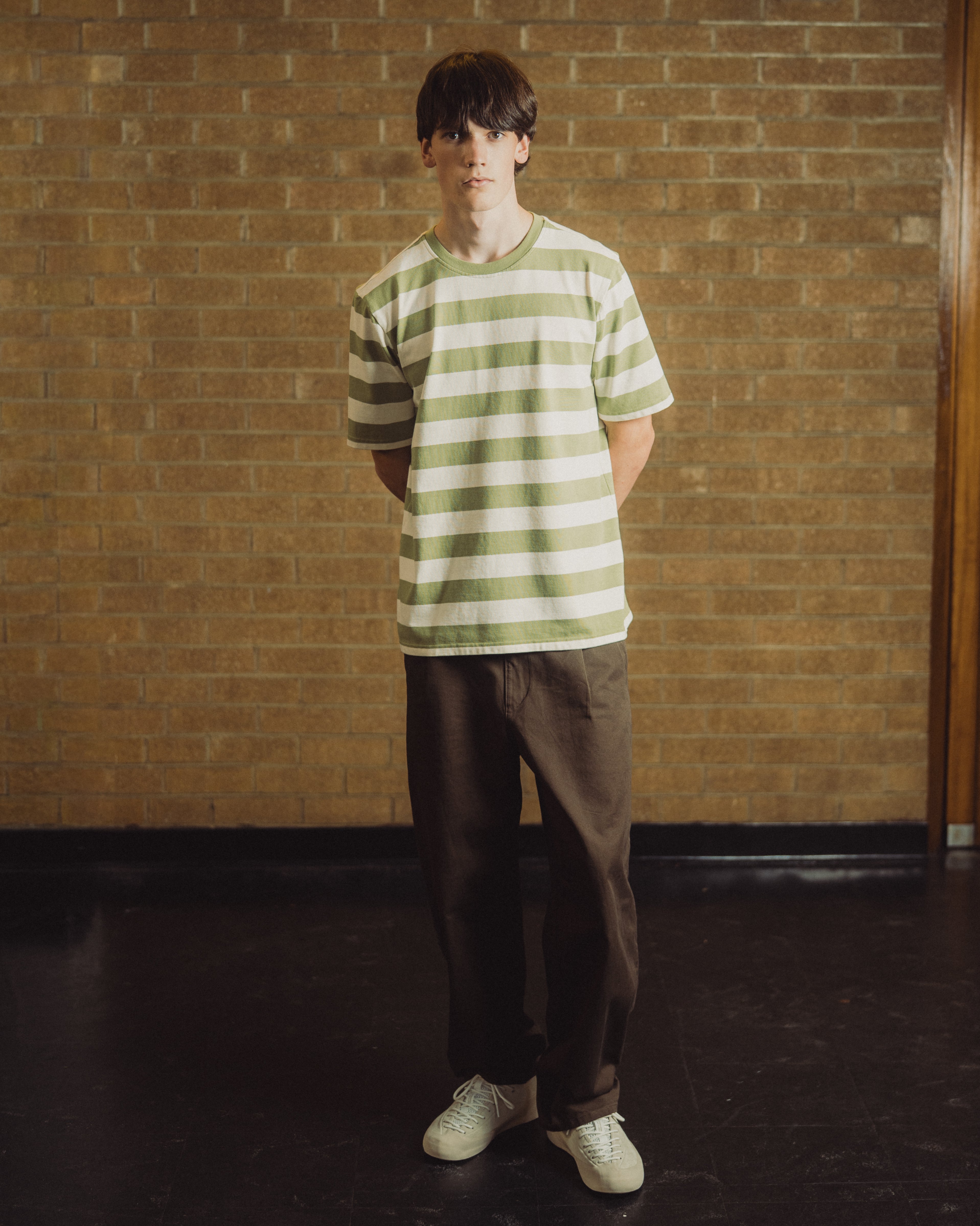 A young man stands in front of a brick wall wearing a light green and white horizontally striped t-shirt, dark brown pants, and white sneakers.